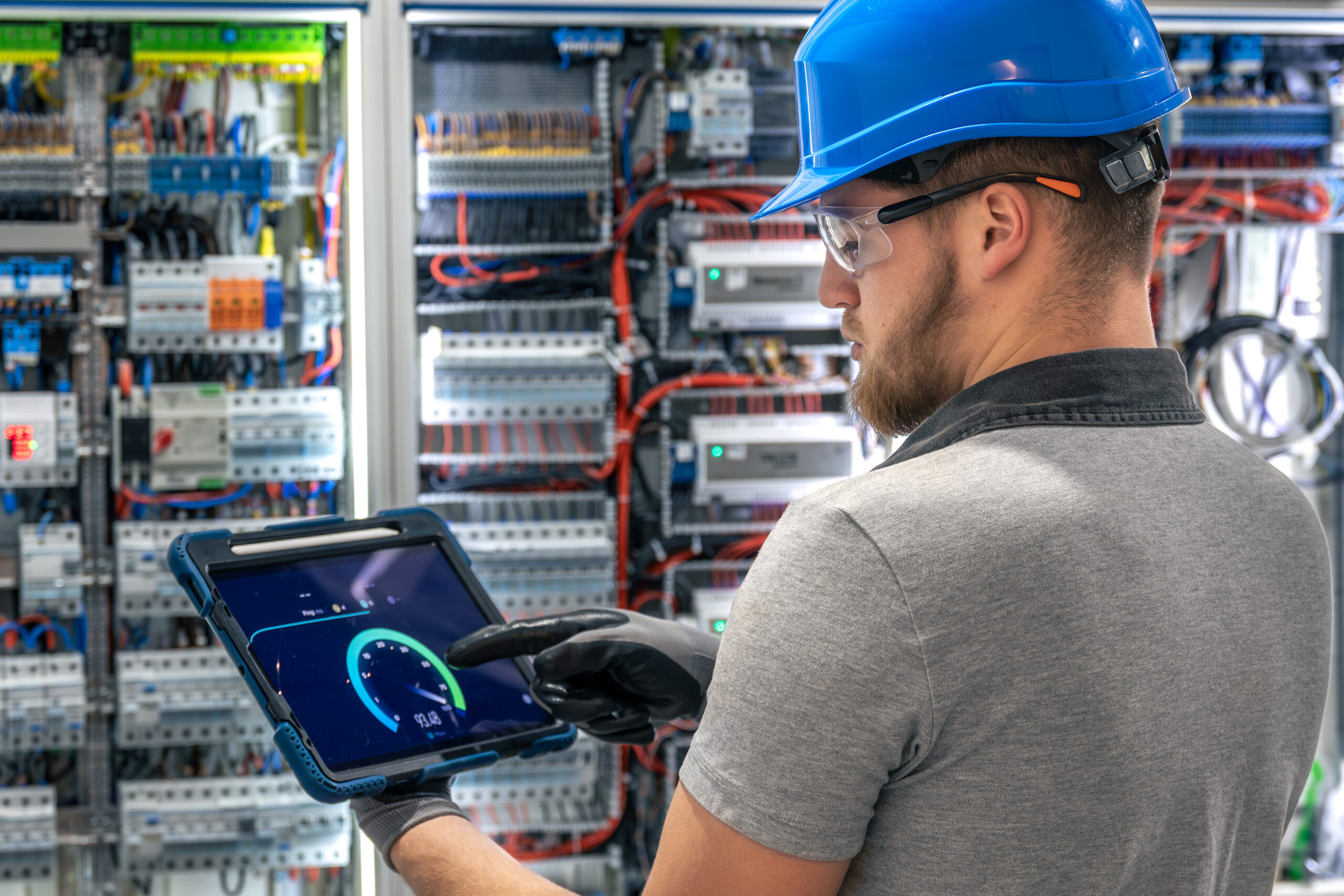 A specialist in helmet and gloves points at a tablet screen while standing before a complex switchboard. Scene of digital diagnostics in an industrial indoor setting.